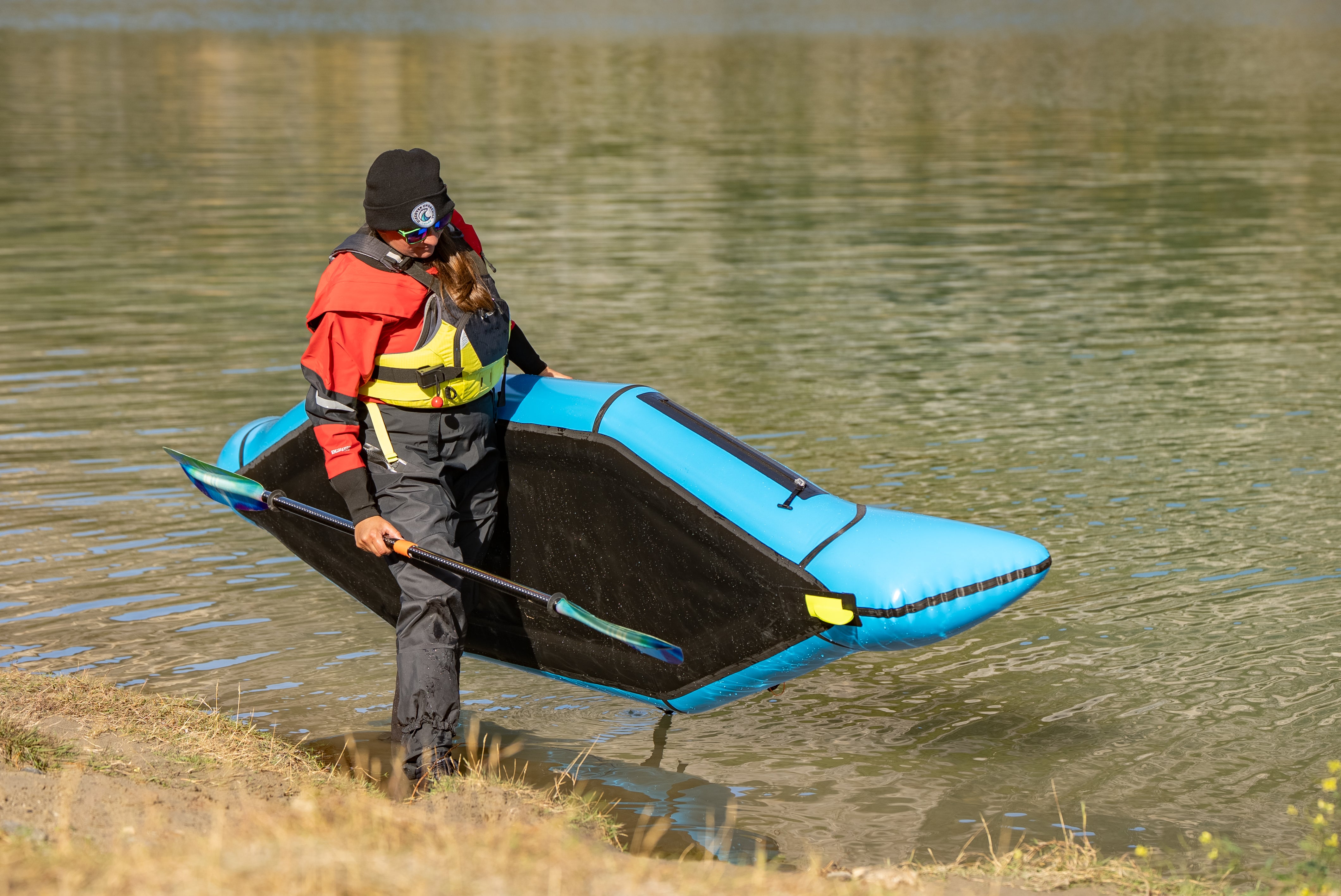 Person carrying a blue inflatable kayak on a grassy bank near water