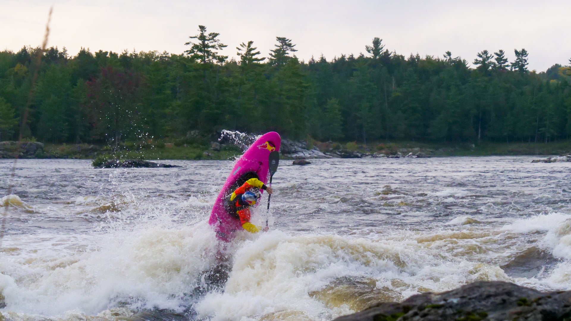 Person in a colorful wetsuit paddling a kayak on turbulent water with a forested shoreline.